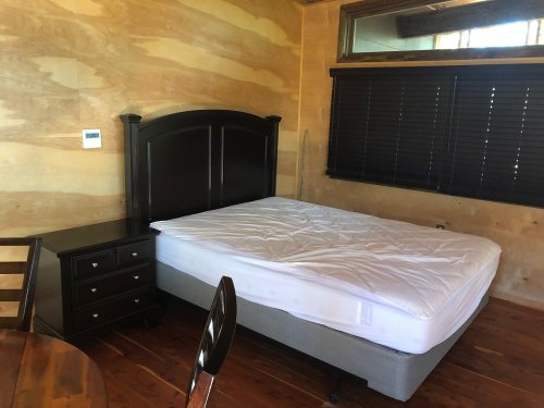 A simple bedroom featuring a wooden bed frame, a mattress, a nightstand, and a dining table with chairs.