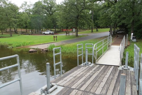 A wooden dock extends over calm water, surrounded by green trees and a grassy area, with a road and parked truck in the background.