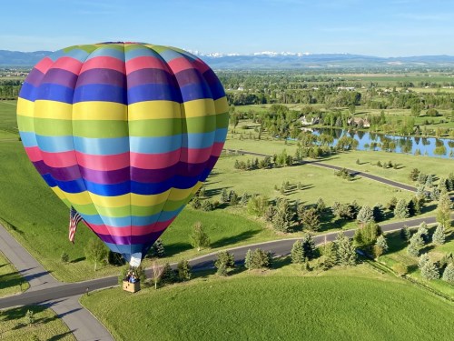 A colorful hot air balloon floats above a green landscape with trees and a pond, under a clear blue sky.