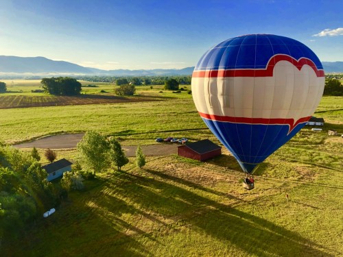 A colorful hot air balloon floats over a green field with mountains in the background and a small building below.