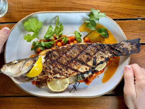 A grilled whole fish garnished with lemon, herbs, and a colorful sauce, served on a white plate on a wooden table.