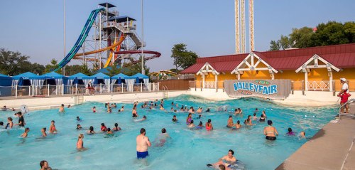 A busy water park scene with people swimming in a pool, colorful slides in the background, and sunny skies.