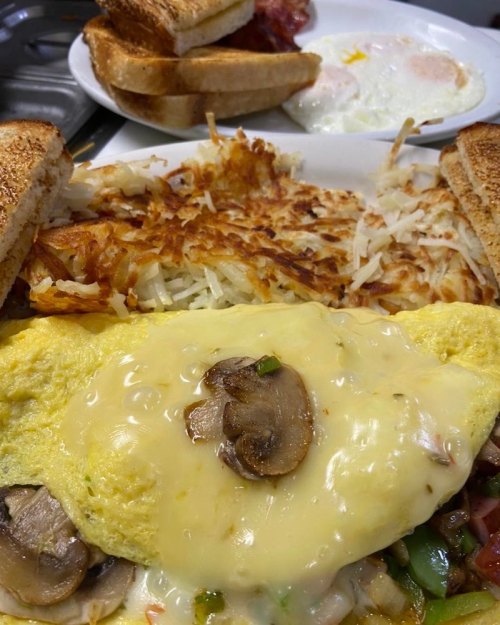 A plate of breakfast featuring a cheesy mushroom omelet, hash browns, toast, and fried eggs in the background.