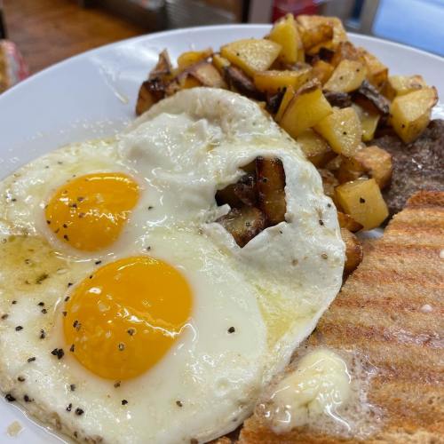 A plate with two sunny-side-up eggs, seasoned potatoes, a slice of toast, and a pat of butter.