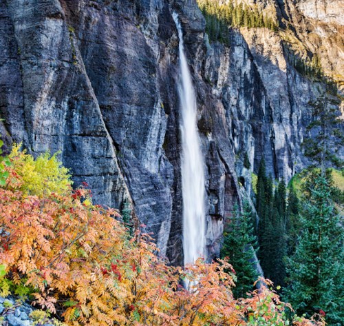 A waterfall cascades down a rocky cliff, surrounded by vibrant autumn foliage and evergreen trees.
