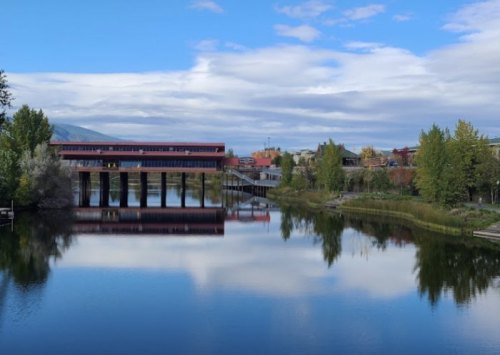 A serene river scene with a modern building on stilts, surrounded by trees and reflecting clouds in the water.