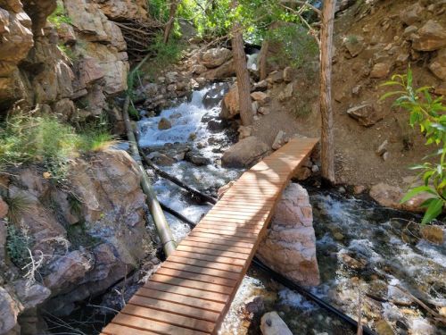 A wooden bridge crosses a rocky stream surrounded by lush greenery and steep stone walls.