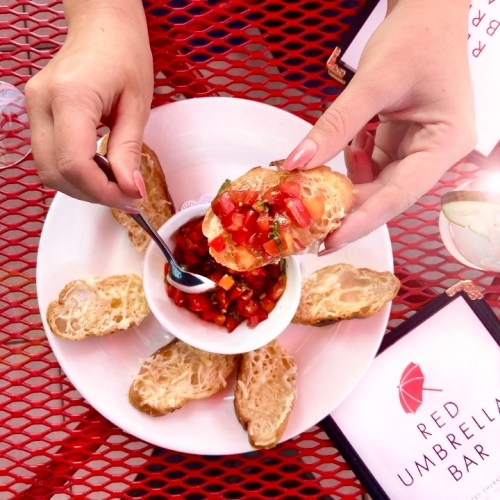 A person holds a spoonful of salsa over a plate of toasted bread slices, with a menu in the background.