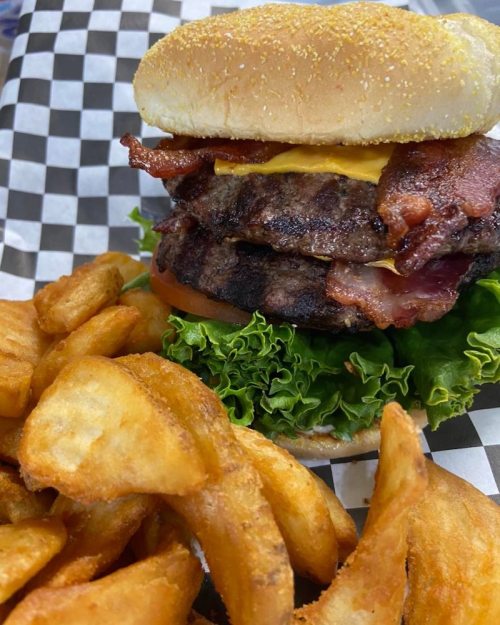 A stacked burger with bacon, cheese, lettuce, and tomato, served with crispy potato wedges on the side.