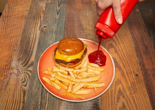 A hand squeezes ketchup onto a cheeseburger and crinkle-cut fries on an orange plate, set on a wooden table.