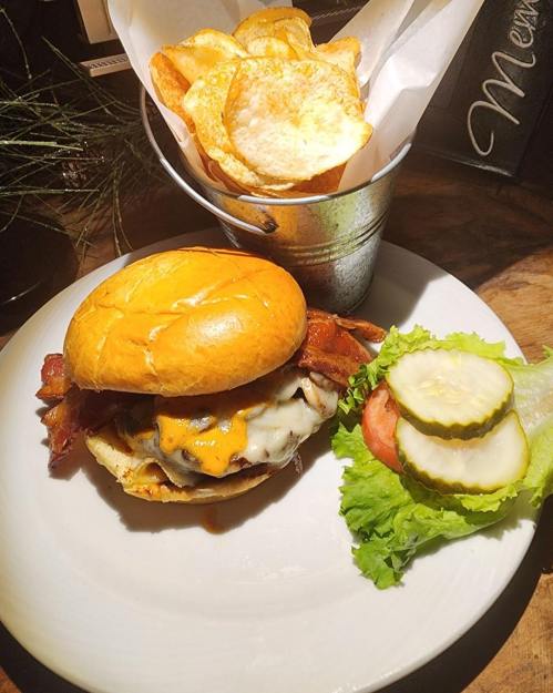 A juicy burger with bacon, cheese, lettuce, tomato, and pickles, served with a side of crispy potato chips in a bucket.