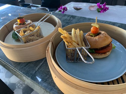Two gourmet burgers with fries served in bamboo steamers, garnished with cherry tomatoes and pickles.