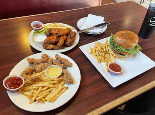 A table with two plates: one with wings and fries, the other with a burger and fries, both with dipping sauces.