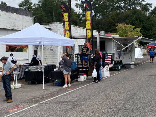A street vendor setup with tents and food trucks, people browsing, and flags displayed along a quiet road.