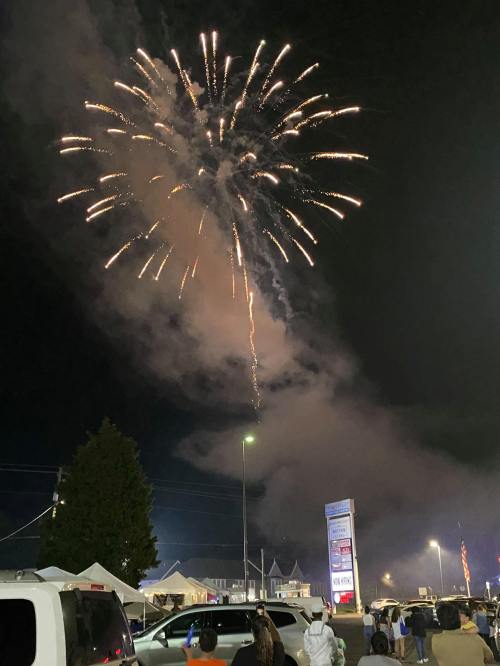 Fireworks burst in the night sky above a crowd, with smoke and cars visible below, creating a festive atmosphere.