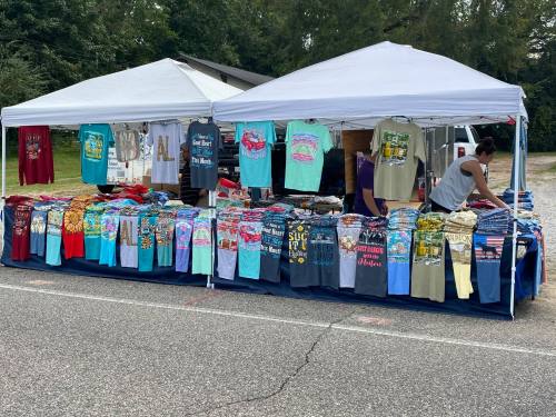 Two tents at a market displaying a variety of colorful t-shirts and clothing items for sale.