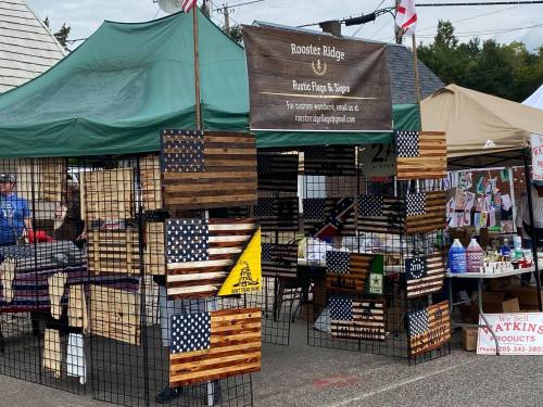 A vendor booth displaying rustic wooden flags and signs, with a sign for "Rooster Ridge" in a market setting.