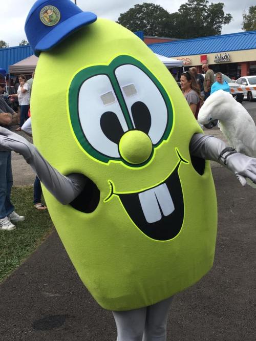 A cheerful green mascot with a big smile and a blue cap stands outdoors, with a parrot perched on its arm.
