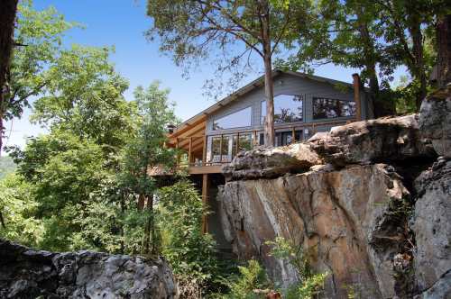 A modern house perched on a rocky ledge, surrounded by lush greenery and trees.