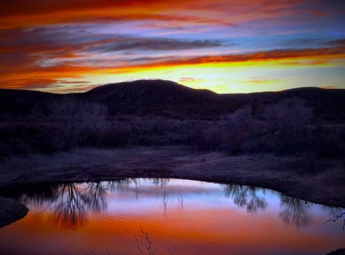A serene sunset over a calm pond, reflecting vibrant orange and blue hues against a backdrop of rolling hills.