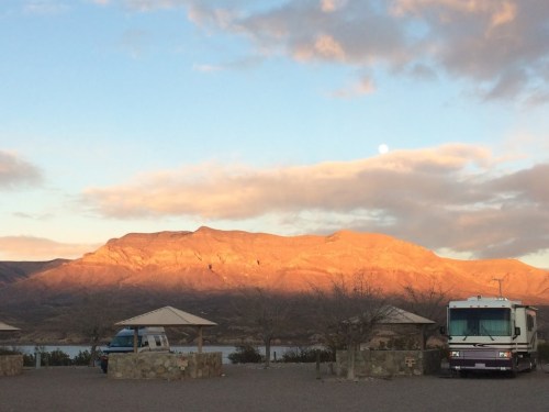 A scenic view of mountains at sunset, with an RV and shaded areas in the foreground and a full moon in the sky.