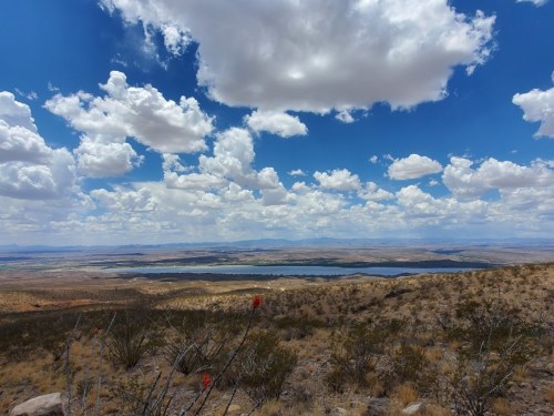 A panoramic view of a desert landscape with a lake, under a bright blue sky filled with fluffy white clouds.