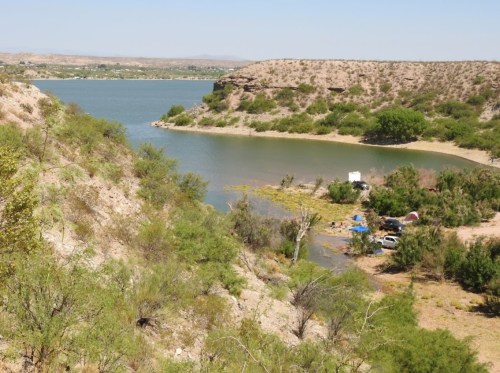 A serene landscape featuring a river winding through hills, with greenery and a small campsite visible along the shore.
