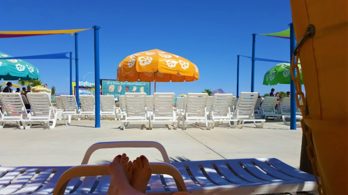 A view from a lounge chair at a beach resort, featuring colorful umbrellas and empty sun loungers under a clear blue sky.