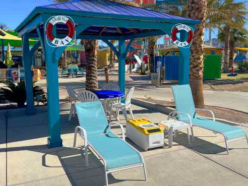 A colorful cabana with lounge chairs and a table, surrounded by palm trees and vibrant umbrellas.