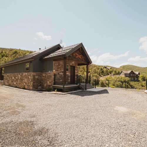 A modern stone and wood house with a porch, surrounded by greenery and mountains under a clear blue sky.