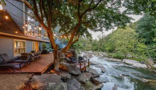 A cozy riverside deck with string lights, surrounded by trees and rocks, near a flowing river and lush greenery.