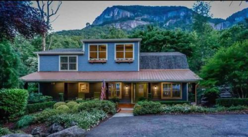 A charming blue house with a porch, surrounded by greenery and mountains in the background, under a twilight sky.