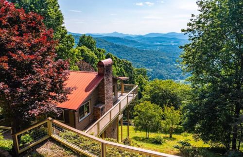 A scenic view of a house on a hillside, surrounded by trees, overlooking rolling mountains and a clear blue sky.