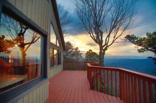 A wooden deck with a view of mountains and a sunset, framed by trees and reflections in the windows of a nearby building.