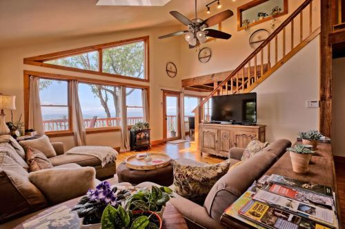 Cozy living room with large windows, a staircase, and a view of nature, featuring comfortable seating and decorative plants.