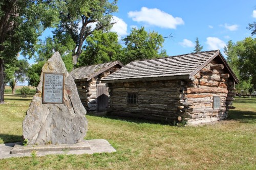 Two rustic log cabins beside a large stone monument in a grassy area under a blue sky with scattered clouds.