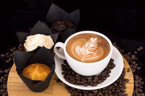 A cup of latte art surrounded by three muffins on a wooden board, with coffee beans scattered around.