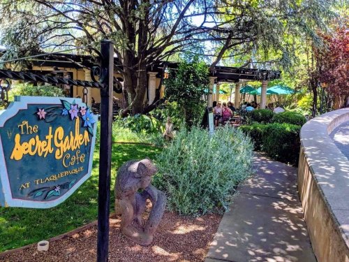 A sign for "The Secret Garden Café" near a lush garden with outdoor seating and a stone pathway.