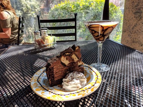 A slice of chocolate cake with whipped cream and a martini with chocolate swirls on a table in a sunny café.