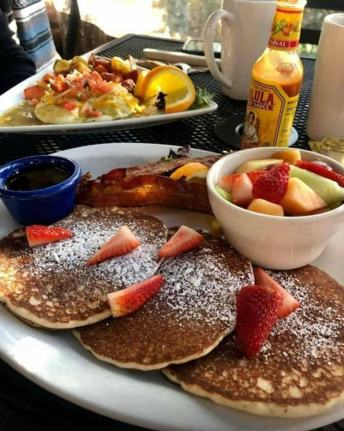 A plate of pancakes topped with strawberries and powdered sugar, alongside a bowl of fruit and a plate of eggs and shrimp.