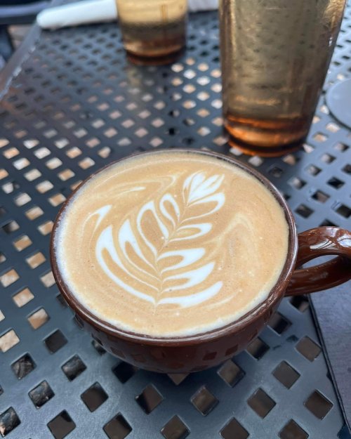 A close-up of a latte in a brown cup, featuring a delicate leaf design in the foam, on a textured black table.