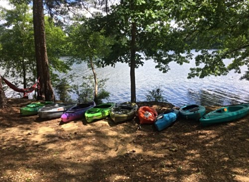 Colorful kayaks lined up on a sandy shore beside a calm lake, surrounded by trees and greenery.