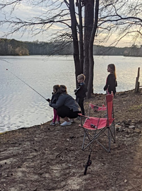 A group of children fishing by a lake, with a pink chair nearby and trees in the background.