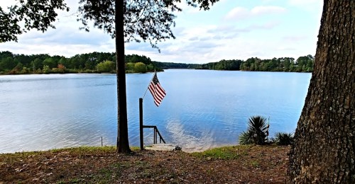 A serene lake view with an American flag hanging by the water, surrounded by trees and a clear blue sky.