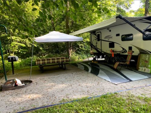 A campsite featuring a parked RV, picnic table, grill, and a shaded area under a canopy surrounded by trees.