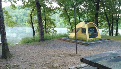 A yellow tent set up by a calm lake, surrounded by trees and a gravel campsite. Mist rises from the water.