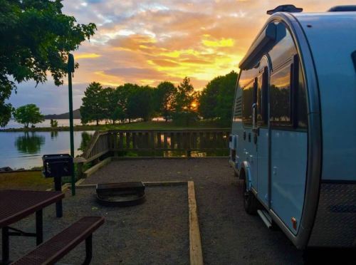 A blue camper parked by a lake at sunset, with trees and a picnic area in the foreground.