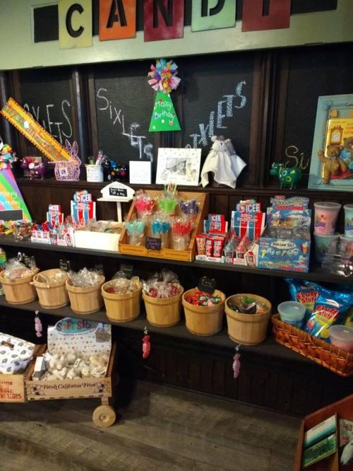 A colorful candy shop display with jars, baskets, and various sweets on wooden shelves. Bright decorations in the background.