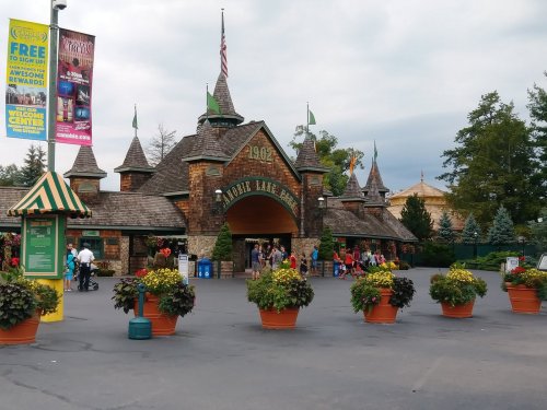 Entrance to a theme park featuring a castle-like structure, surrounded by large flower pots and visitors.