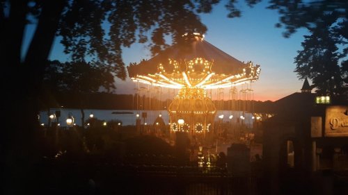 A brightly lit carousel at dusk, surrounded by trees and a lake, with a colorful sunset in the background.